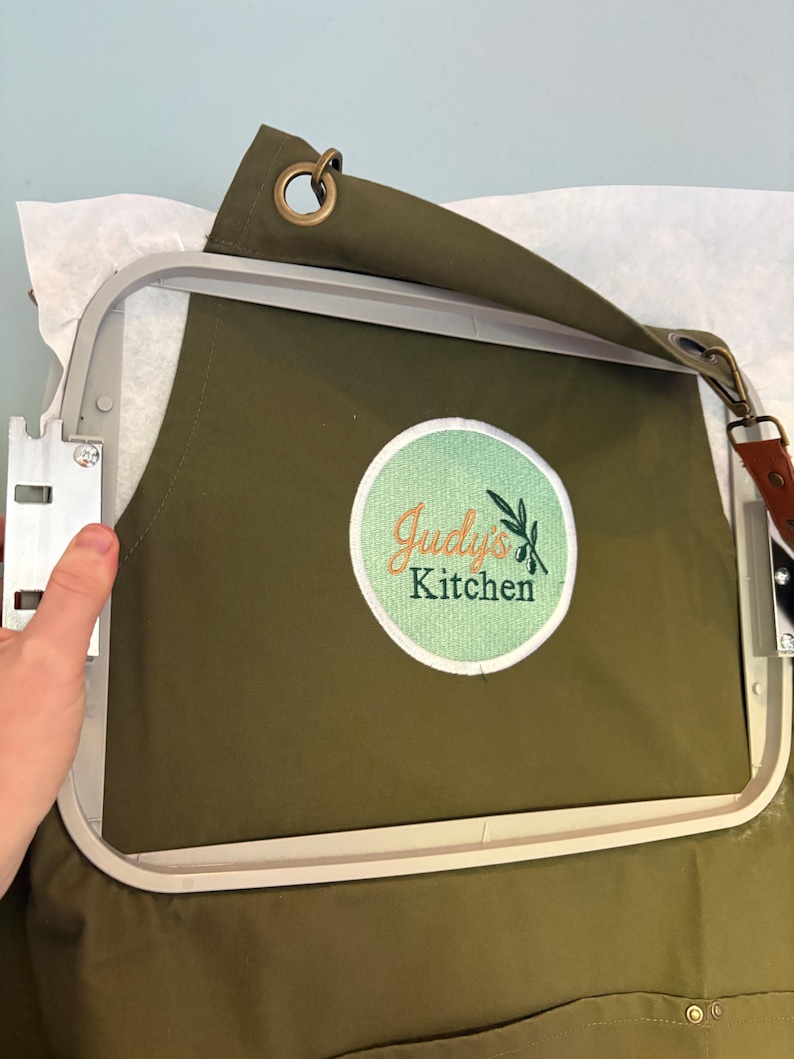 May include: Olive green apron with a white and orange embroidered logo that reads "Judy's Kitchen" with olive branch accents. The apron is in an embroidery hoop, ready for stitching. Features a brown leather strap and metal grommets.