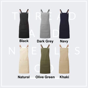 May include: Aprons in six colors: black, dark gray, navy, natural, olive green, and khaki. Each apron has a large front pocket and adjustable straps with leather-like accents. The aprons are labeled with their respective colors.