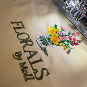 May include: Embroidered design featuring a floral arrangement in a vase, with yellow sunflowers, pink and red flowers, and green leaves. The text "FLORALS By Moll" is embroidered in a dark green color on a beige fabric.