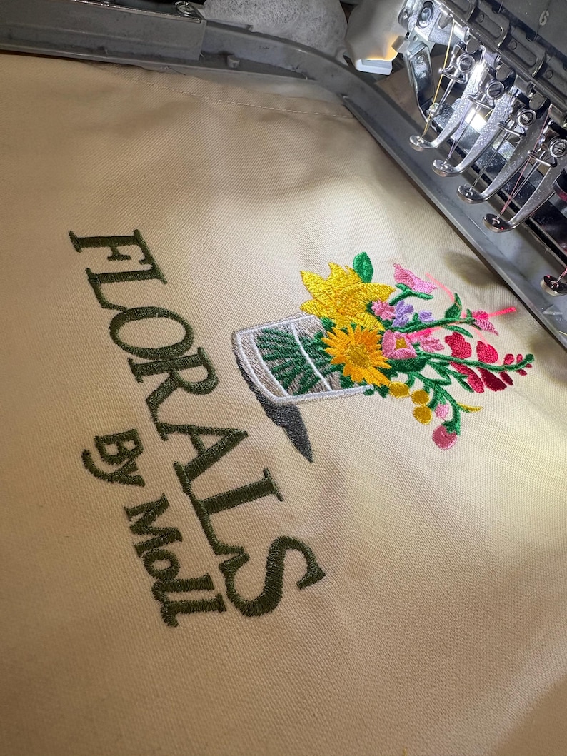 May include: Embroidered design featuring a floral arrangement in a vase, with yellow sunflowers, pink and red flowers, and green leaves. The text "FLORALS By Moll" is embroidered in a dark green color on a beige fabric.
