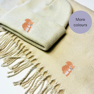Pode incluir: Um gorro de malha bege e uma echarpe combinando com um esquilo castanho bordado em cada um. O texto "Mais cores" está em um círculo roxo no canto superior direito da imagem.