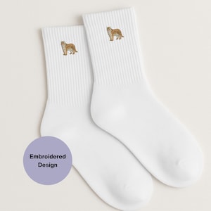 Puede incluir: Dos calcetines blancos con puños acanalados, cada uno bordado con un diseño detallado de tigre en tonos marrones y beige. Los calcetines se muestran sobre un fondo claro. Un círculo morado con el texto "Embroidered Design" está en la parte inferior izquierda.
