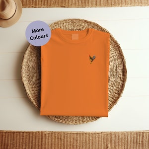 May include: Orange t-shirt with a small embroidered rooster design. The t-shirt is folded and placed on a woven basket.