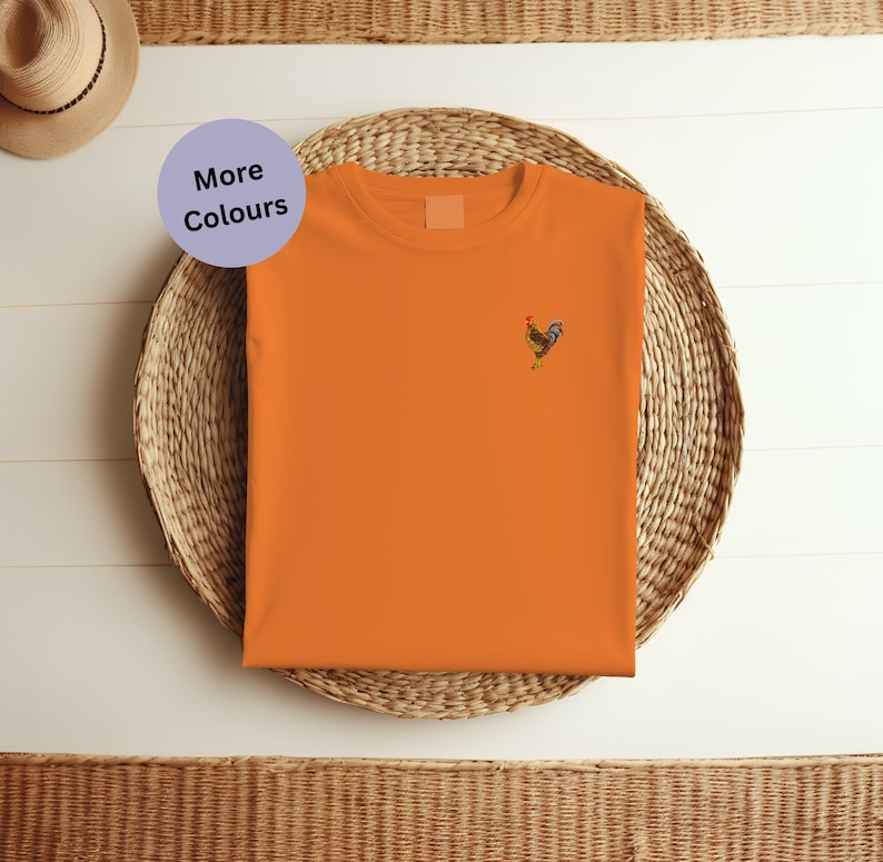 May include: Orange t-shirt with a small embroidered rooster design. The t-shirt is folded and placed on a woven basket.