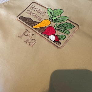 May include: Tan apron with an embroidered design featuring a carrot, radish, and beet with green leaves, and the words "HOME GROWN" and "Pia". The apron is made of a durable fabric.