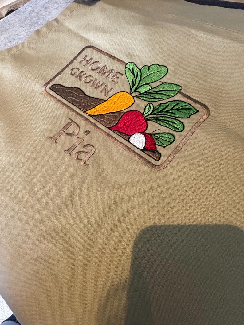 May include: Tan apron with an embroidered design featuring a carrot, radish, and beet with green leaves, and the words "HOME GROWN" and "Pia". The apron is made of a durable fabric.