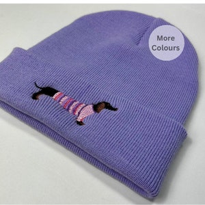 May include: Purple knitted beanie with a dachshund wearing a pink and purple striped sweater embroidered on the front. The beanie has the text "More Colours" on the side.