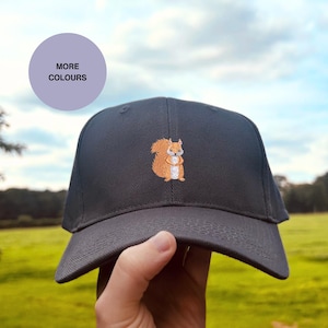 Könnte beinhalten: Dunkelgraue Baseballkappe mit einem gestickten orangefarbenen Eichhörnchen. Die Kappe wird vor dem Hintergrund einer grünen Wiese und eines blauen Himmels gehalten. Ein violetter Kreis mit der Aufschrift "MORE COLOURS" ist im Hintergrund zu sehen.
