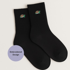 May include: Two black crew socks, each featuring a small embroidered turtle motif in green, brown, and white. The socks are made from a ribbed knit fabric. A purple circle with the text "Embroidered Design" is in the lower left corner.