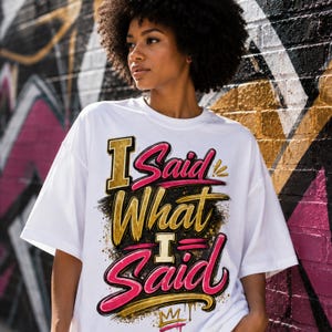 May include: A white short-sleeved t-shirt featuring a graphic print with the text I Said What I Said in stylised pink and gold lettering. The design includes black brushstroke accents, gold paint splatters, and a small gold crown icon at the base.