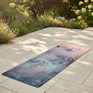 May include: A rectangular yoga mat featuring a mottled, abstract pattern in shades of deep blue, slate grey, and soft peach. The surface has a textured appearance with blended colour transitions, laid flat on a light-coloured stone patio surface.