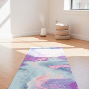 May include: A yoga mat with a marbled pattern in purple, pink, and blue tones is laid out on a wooden floor. The mat is rectangular and made of a flexible material. A diffuser and stacked cushions are visible in the background, in a well-lit room.