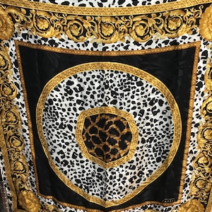 May include: A square silk scarf with a black, white, and gold design. The scarf features a central leopard print pattern surrounded by a black border and ornate gold scrollwork. The edges have a repeating pattern of black and white spots.