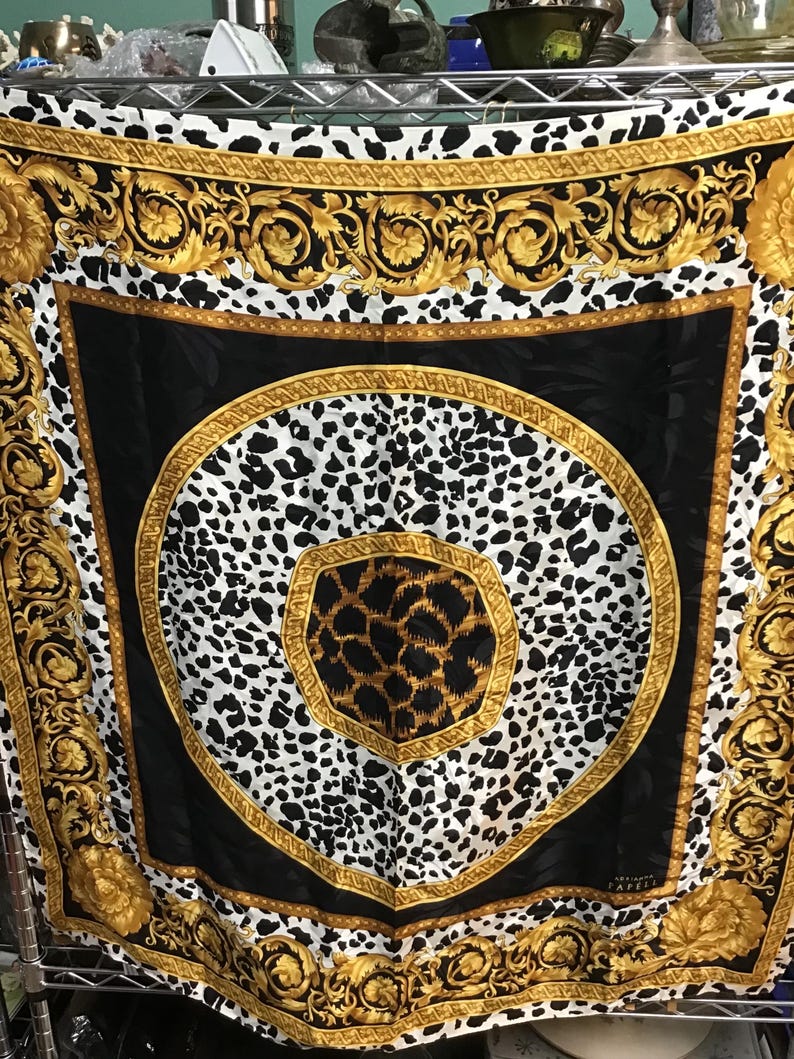 May include: A square silk scarf with a black, white, and gold design. The scarf features a central leopard print pattern surrounded by a black border and ornate gold scrollwork. The edges have a repeating pattern of black and white spots.