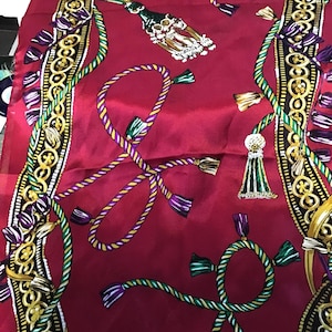 May include: A red scarf with a repeating pattern of ropes, tassels, and decorative borders. The ropes are in shades of purple, yellow, and green. The tassels are gold, green, and white. The borders are gold and black.