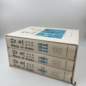 May include: A set of three books titled "WEEDS OF KOREA" in a light beige slipcase. The spines display the title in English and Korean. The top of the slipcase features a blue and white image. The books are stacked.