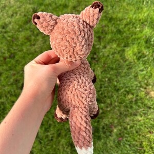 May include: A handmade, plush fox toy in shades of brown and white. The fox has a textured, fuzzy appearance, with darker brown accents on the ears and paws. The tail is long and fluffy, with a white tip. The toy is held against a background of green grass.