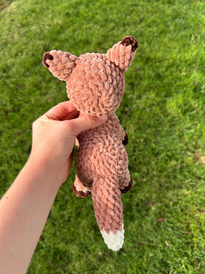 May include: A handmade, plush fox toy in shades of brown and white. The fox has a textured, fuzzy appearance, with darker brown accents on the ears and paws. The tail is long and fluffy, with a white tip. The toy is held against a background of green grass.