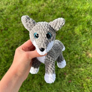May include: A gray and white crocheted wolf plush toy with blue eyes and a black nose. The handmade stuffed animal has a fluffy texture and is held against a green background.