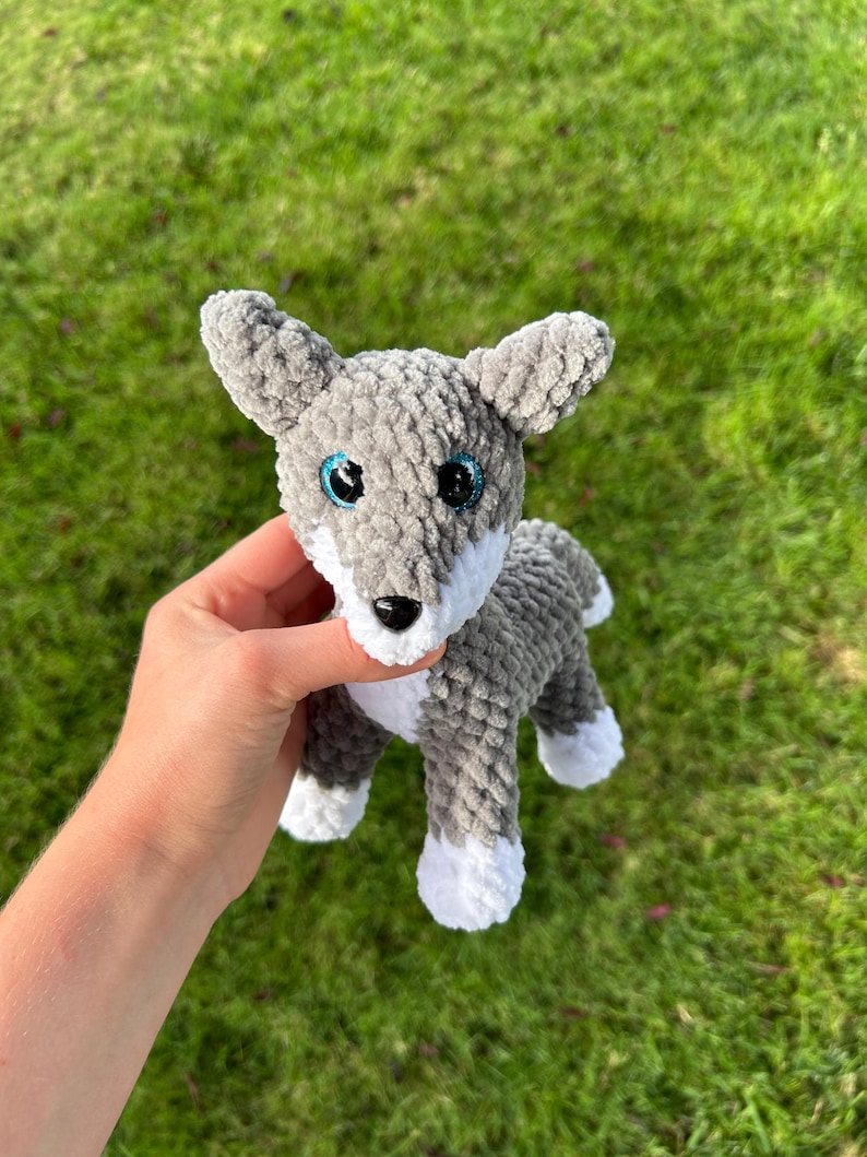 May include: A gray and white crocheted wolf plush toy with blue eyes and a black nose. The handmade stuffed animal has a fluffy texture and is held against a green background.