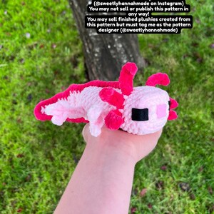 Op de afbeelding: Een handgemaakte gehaakte axolotl-knuffel in roze en wit, met felroze kieuwen en accenten. De knuffel wordt in een hand gehouden tegen een achtergrond van groen gras en een boomstam. Tekst op de afbeelding geeft de patroonontwerper aan.