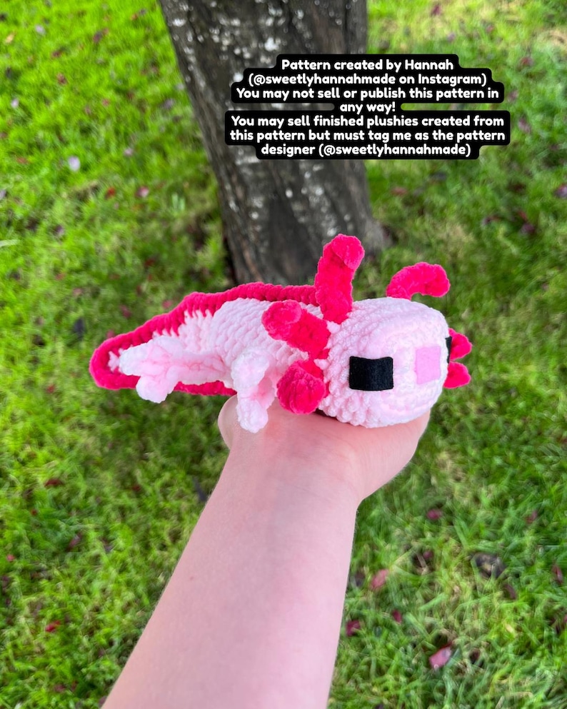 Op de afbeelding: Een handgemaakte gehaakte axolotl-knuffel in roze en wit, met felroze kieuwen en accenten. De knuffel wordt in een hand gehouden tegen een achtergrond van groen gras en een boomstam. Tekst op de afbeelding geeft de patroonontwerper aan.