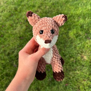 May include: A crocheted fox plushie in shades of brown, tan, and white. The fox has dark brown accents on its ears, paws, and nose, with black safety eyes. The plush toy is held against a background of green grass.