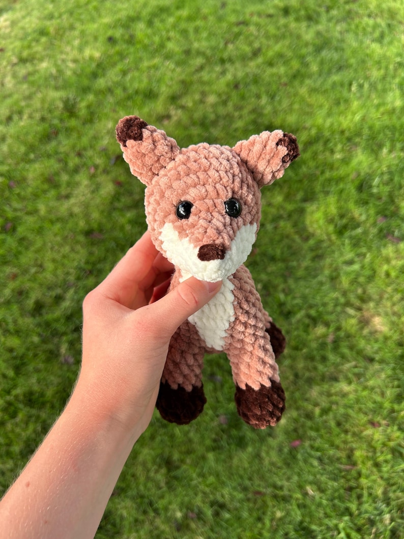 May include: A crocheted fox plushie in shades of brown, tan, and white. The fox has dark brown accents on its ears, paws, and nose, with black safety eyes. The plush toy is held against a background of green grass.
