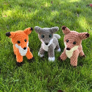 May include: Three crocheted fox and wolf stuffed animals in orange, gray, and brown. The fox has black ears and feet, the wolf has gray ears and feet, and the brown fox has brown ears and feet. All have white accents and black eyes.