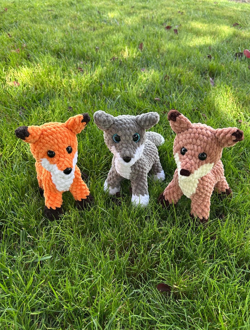 May include: Three crocheted fox and wolf stuffed animals in orange, gray, and brown. The fox has black ears and feet, the wolf has gray ears and feet, and the brown fox has brown ears and feet. All have white accents and black eyes.