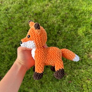 May include: A handmade orange and white plush fox toy with brown accents. The fox has a fluffy texture, black eyes, and a white chest and tail tip. The fox is held against a background of green grass.