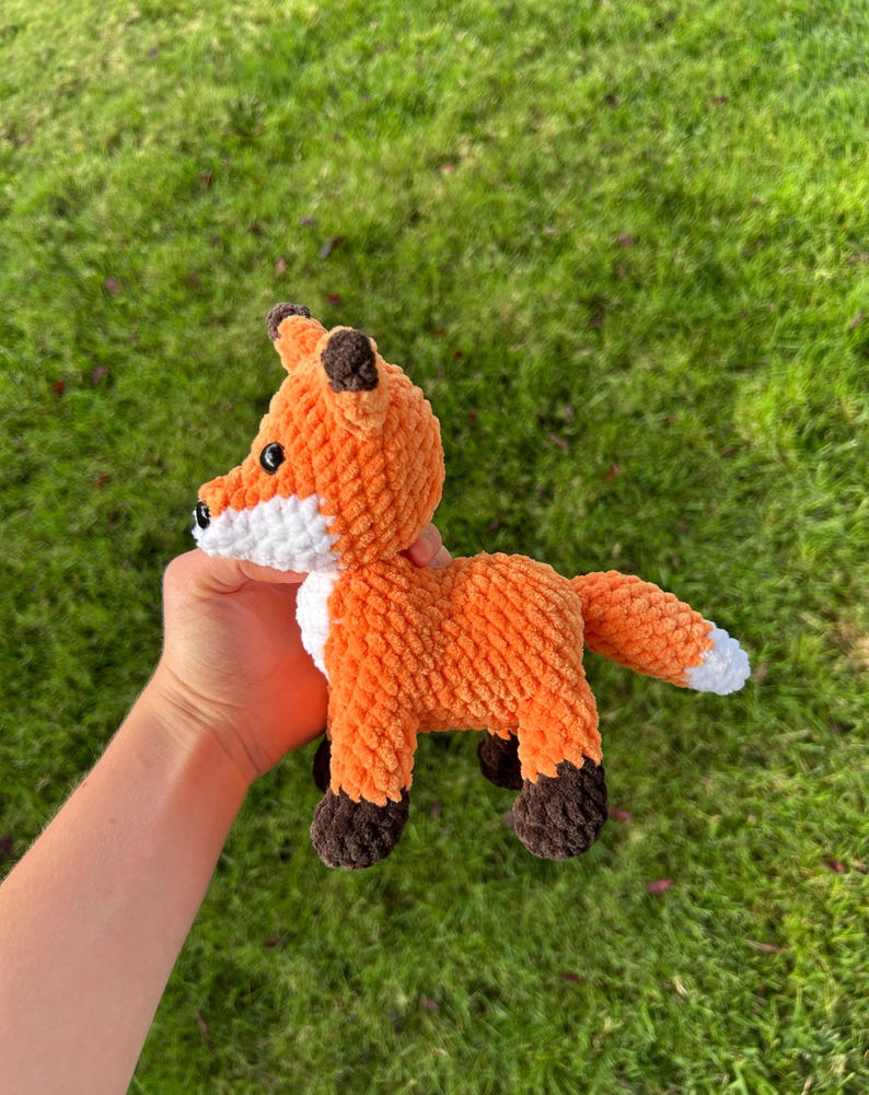May include: A handmade orange and white plush fox toy with brown accents. The fox has a fluffy texture, black eyes, and a white chest and tail tip. The fox is held against a background of green grass.