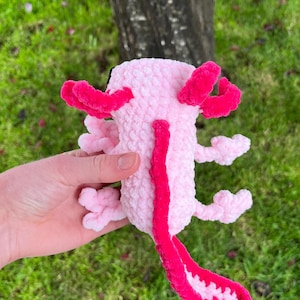 Op de afbeelding: Een roze axolotl knuffel met felroze kieuwen en een staart. Het speelgoed is gemaakt van zacht, getextureerd materiaal. De axolotl wordt in een hand gehouden tegen een achtergrond van groen gras en een boomstam. De knuffel heeft een gedetailleerd ontwerp.