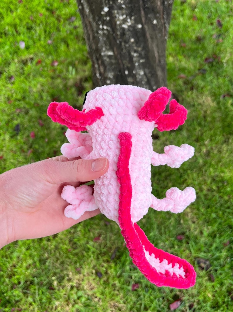 Op de afbeelding: Een roze axolotl knuffel met felroze kieuwen en een staart. Het speelgoed is gemaakt van zacht, getextureerd materiaal. De axolotl wordt in een hand gehouden tegen een achtergrond van groen gras en een boomstam. De knuffel heeft een gedetailleerd ontwerp.