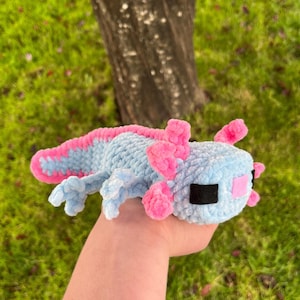Op de afbeelding: Een gehaakte axolotl-knuffel in lichtblauw met roze kieuwen en accenten. Het handgemaakte speelgoed heeft zwarte ogen en een roze mond. De axolotl ligt op een groene ondergrond.