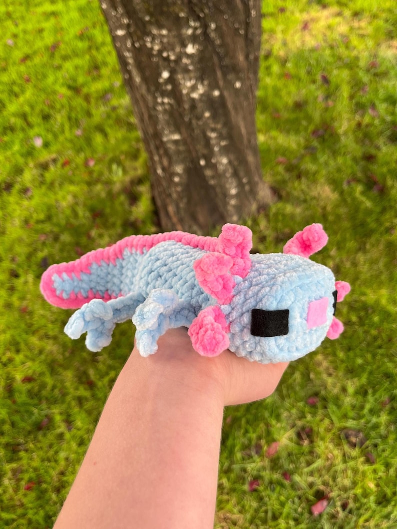 Op de afbeelding: Een gehaakte axolotl-knuffel in lichtblauw met roze kieuwen en accenten. Het handgemaakte speelgoed heeft zwarte ogen en een roze mond. De axolotl ligt op een groene ondergrond.