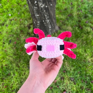 Op de afbeelding: Een handgemaakt, pluche axolotl-speeltje in roze en wit. De axolotl heeft felroze kieuwen en een roze mond. De ogen zijn zwart en het lichaam is zacht, lichtroze. Het speelgoed is gemaakt van een zacht materiaal.