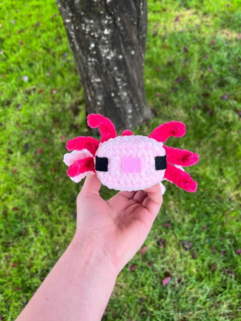 Op de afbeelding: Een handgemaakt, pluche axolotl-speeltje in roze en wit. De axolotl heeft felroze kieuwen en een roze mond. De ogen zijn zwart en het lichaam is zacht, lichtroze. Het speelgoed is gemaakt van een zacht materiaal.