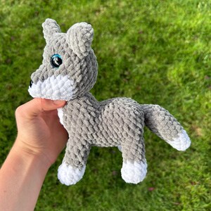 May include: A gray and white crocheted wolf plush toy with blue eyes and a black nose. The handmade stuffed animal has a fluffy texture and is standing on all fours. The wolf's paws and snout are white.