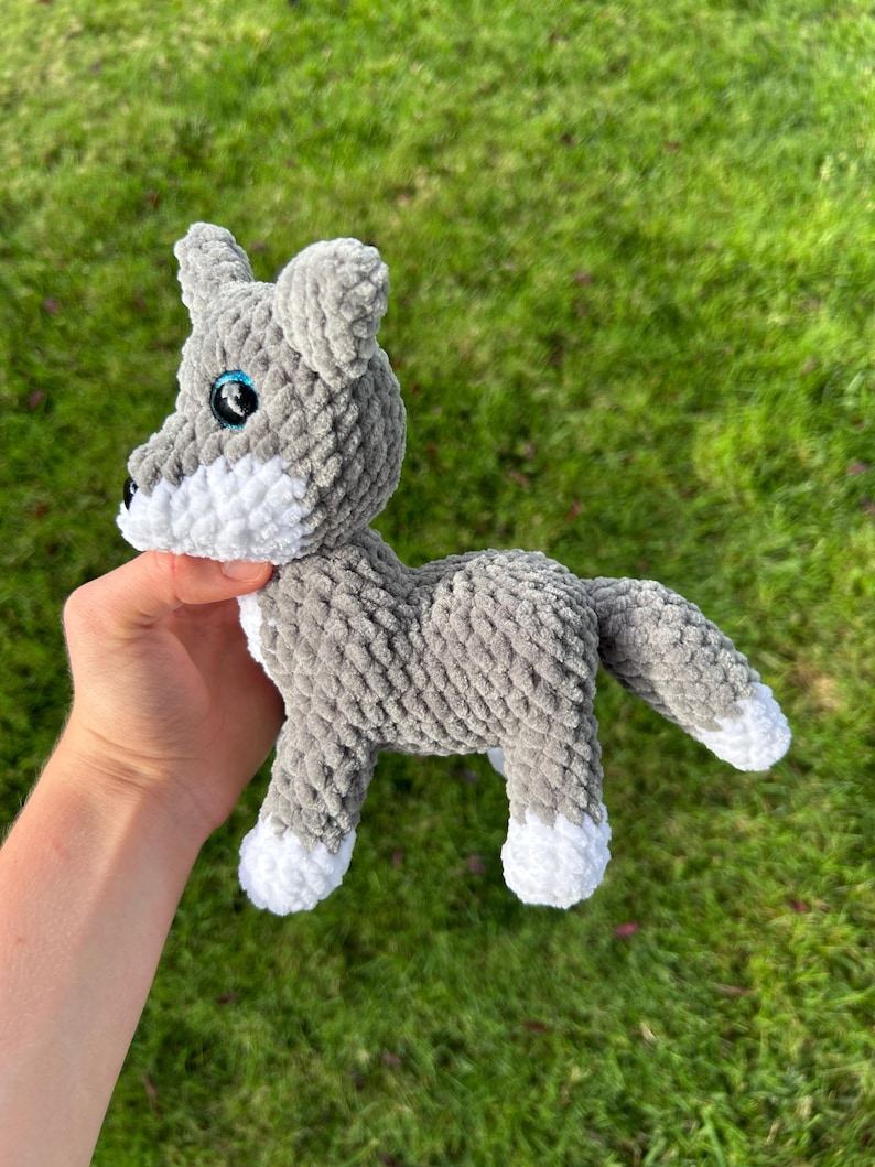 May include: A gray and white crocheted wolf plush toy with blue eyes and a black nose. The handmade stuffed animal has a fluffy texture and is standing on all fours. The wolf's paws and snout are white.