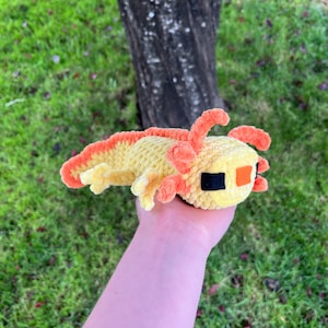Op de afbeelding: Een handgemaakt, pluche axolotl-speeltje in geel en oranje tinten. Het speelgoed heeft een gele body met oranje kieuwen en een rugvin. De axolotl heeft zwarte en oranje ogen. Het speelgoed wordt in een hand gehouden tegen een achtergrond van groen gras en een boomstam.