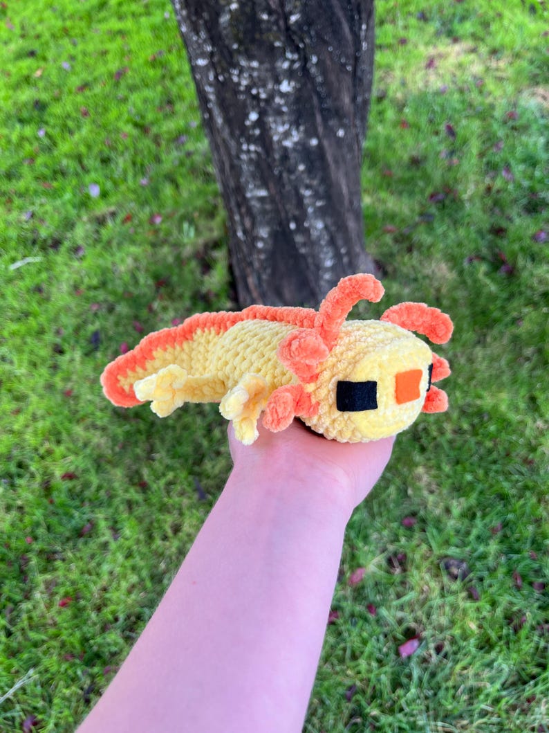 Op de afbeelding: Een handgemaakt, pluche axolotl-speeltje in geel en oranje tinten. Het speelgoed heeft een gele body met oranje kieuwen en een rugvin. De axolotl heeft zwarte en oranje ogen. Het speelgoed wordt in een hand gehouden tegen een achtergrond van groen gras en een boomstam.