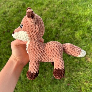 May include: A crocheted fox toy in shades of brown, white, and dark brown. The plush fox has a textured, soft appearance, with a white chest and muzzle, and dark brown accents on the ears and feet. The fox is posed on a green grassy background.