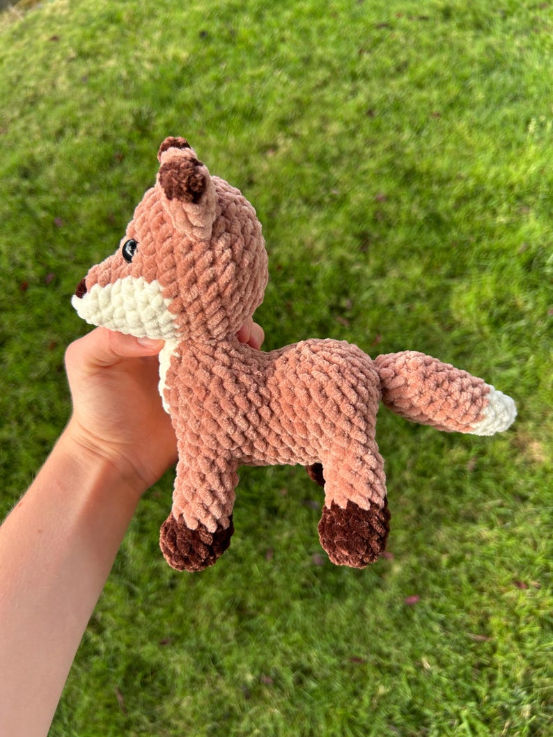 May include: A crocheted fox toy in shades of brown, white, and dark brown. The plush fox has a textured, soft appearance, with a white chest and muzzle, and dark brown accents on the ears and feet. The fox is posed on a green grassy background.
