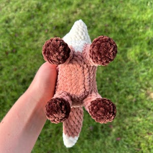 May include: A handmade crochet fox toy, viewed from below, with a white and pink body, brown paws, and a white-tipped tail. The fox is made of soft, textured yarn, perfect for a child's toy or nursery decor.