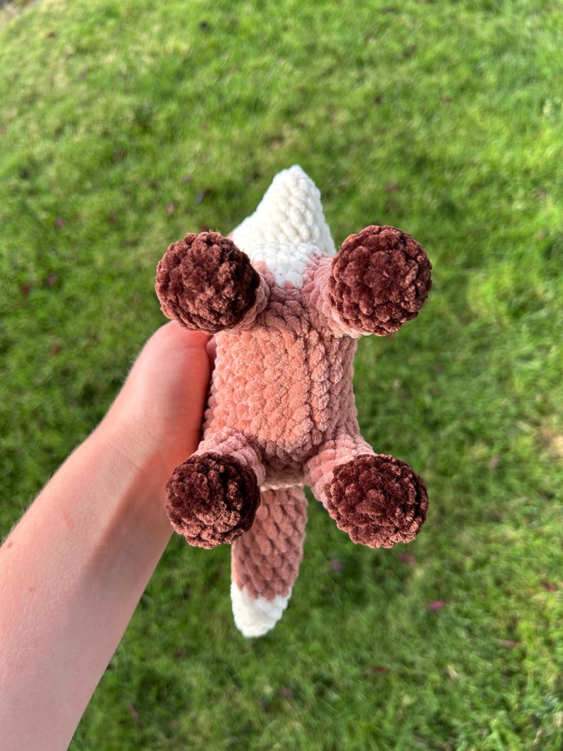 May include: A handmade crochet fox toy, viewed from below, with a white and pink body, brown paws, and a white-tipped tail. The fox is made of soft, textured yarn, perfect for a child's toy or nursery decor.