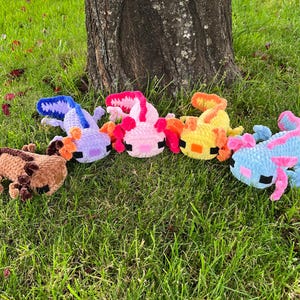 Op de afbeelding: Een verzameling kleurrijke, gehaakte axolotl-knuffels in verschillende kleuren, waaronder bruin, paars, roze, oranje, geel en blauw. De knuffels zijn gerangschikt op groen gras bij een boomstam.