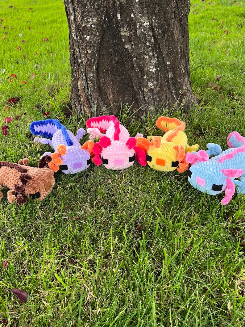 Op de afbeelding: Een verzameling kleurrijke, gehaakte axolotl-knuffels in verschillende kleuren, waaronder bruin, paars, roze, oranje, geel en blauw. De knuffels zijn gerangschikt op groen gras bij een boomstam.
