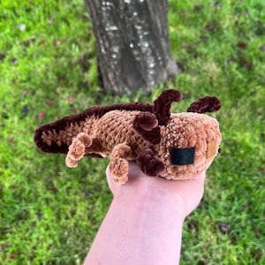 Op de afbeelding: Een handgemaakt, gehaakt axolotl-knuffel in bruin- en bruintinten. Het gehaakte wezen heeft donkerbruine kieuwen, een staart en kleine, vierkante zwarte ogen. Het speelgoed wordt in een hand gehouden tegen een achtergrond van groen gras en een boomstam.