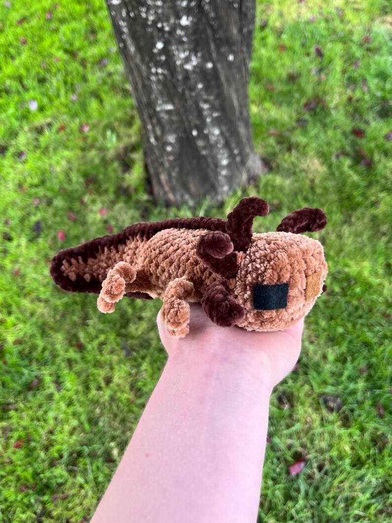 Op de afbeelding: Een handgemaakt, gehaakt axolotl-knuffel in bruin- en bruintinten. Het gehaakte wezen heeft donkerbruine kieuwen, een staart en kleine, vierkante zwarte ogen. Het speelgoed wordt in een hand gehouden tegen een achtergrond van groen gras en een boomstam.